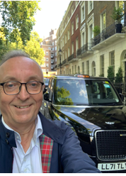 Person taking a selfie with a black taxi on a street lined with trees and buildings.